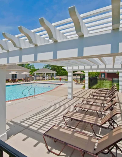 A view from underneath a white wooden pergola showing rows of lounge chairs on a concrete sundeck next to a swimming pool